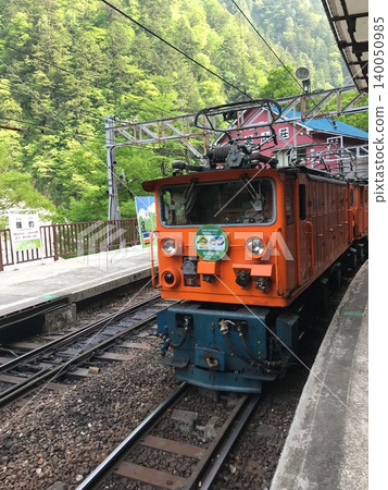An orange electric locomotive and a trolley train stopped at Kanetsuri Station on the Kurobe Gorge Railway. An orange electric locomotive and a trolley train stopped at Kanetsuri Station on the Kurobe Gorge Railway. 140050985