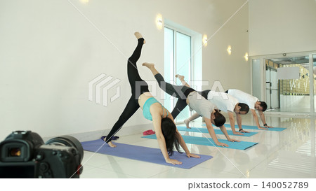 Group of people practicing three legged downward dog yoga pose in modern studio, lifting one leg to build strength, improve balance and flexibility during dynamic fitness and wellness training session Group of people practicing three legged downward dog yoga pose in modern studio, lifting one leg to build strength, improve balance and flexibility during dynamic fitness and wellness training session 140052789