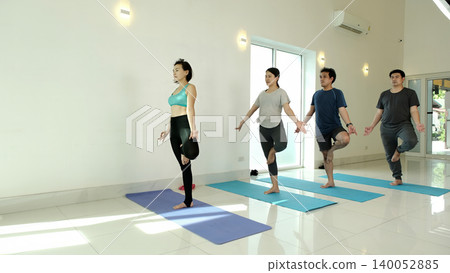 Yoga instructor leading group in tree pose balance exercise in bright studio, focusing on stability, concentration and body alignment to improve core strength and mindfulness during class practice 140052885