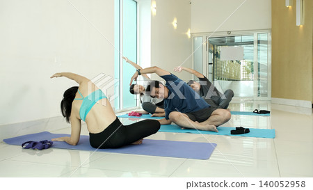 Group of people practicing seated side stretch and lotus yoga pose in bright home studio, improving flexibility, spine mobility and relaxation during guided wellness workout session with instructor Group of people practicing seated side stretch and lotus yoga pose in bright home studio, improving flexibility, spine mobility and relaxation during guided wellness workout session with instructor 140052958