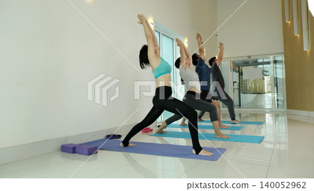 Group of people practicing warrior yoga pose in bright modern studio, stretching legs and opening chest to improve balance, strength and flexibility during guided fitness and wellness class Group of people practicing warrior yoga pose in bright modern studio, stretching legs and opening chest to improve balance, strength and flexibility during guided fitness and wellness class 140052962
