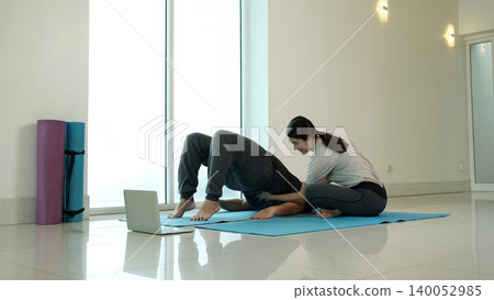 Woman assisting man in bridge yoga pose during home workout session, improving flexibility, core strength and posture with guided support, in bright minimalist fitness studio with laptop training 140052985