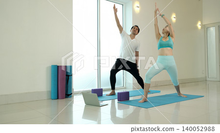 Couple practicing warrior yoga pose with raised arms at home using laptop guidance, improving balance, flexibility and strength, promoting healthy active lifestyle in bright minimalist indoor workout Couple practicing warrior yoga pose with raised arms at home using laptop guidance, improving balance, flexibility and strength, promoting healthy active lifestyle in bright minimalist indoor workout 140052988