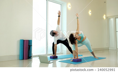 Couple practicing triangle yoga pose with blocks at home using laptop guidance, improving flexibility, balance and posture, strengthening core and legs, promoting healthy active lifestyle in bright 140052991