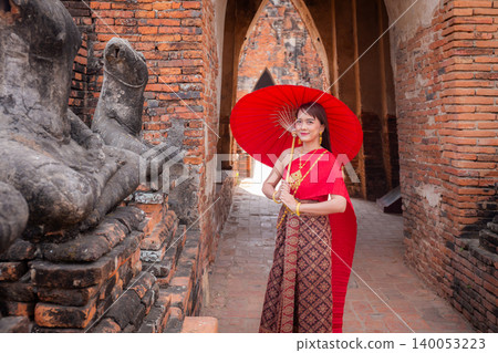 young woman wearing traditional red Thai dress and golden accessories stands holding a traditional umbrella in the historical site Wat Chaiwatthanaram temple. Thai national costume young woman wearing traditional red Thai dress and golden accessories stands holding a traditional umbrella in the historical site Wat Chaiwatthanaram temple. Thai national costume 140053223