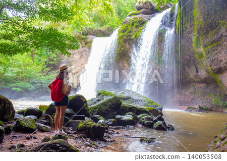 Female tourist wearing a hat and carrying an adventure backpack stands in front of Heo Suwat Waterfall, Khao Yai National Park, Nakhon Ratchasima, Thailand. 140053508