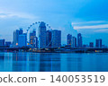 A modern building with a giant Ferris wheel by the sea, at twilight, overlooking the Singapore skyline. 140053519
