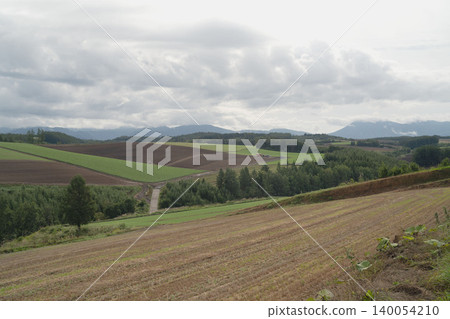 The patchwork hills and farm roads of Biei, Hokkaido, with the distant view of the Tokachi Mountain Range, a cloudy rural landscape in early autumn in September. The patchwork hills and farm roads of Biei, Hokkaido, with the distant view of the Tokachi Mountain Range, a cloudy rural landscape in early autumn in September. 140054210