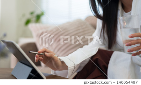 Asian women drinking water holding glass using digital tablet stylus pen writing morning routine healthy lifestyle wellness habit at home living room sofa 140054497
