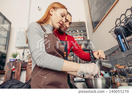 A young Caucasian woman and a young Middle-Eastern man prepare coffee in a cozy cafe. They are focused on their task, surrounded by coffee-making equipment. A young Caucasian woman and a young Middle-Eastern man prepare coffee in a cozy cafe. They are focused on their task, surrounded by coffee-making equipment. 140055270