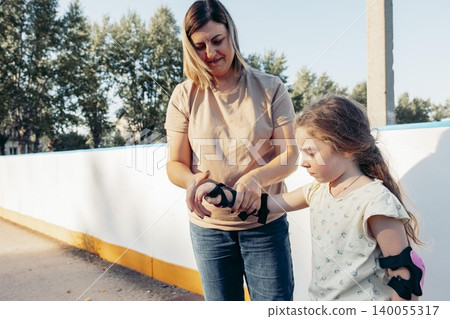 woman helps young girl put on roller skate protective gear. They are outdoors, enjoying quality time together. family bonding and activity woman helps young girl put on roller skate protective gear. They are outdoors, enjoying quality time together. family bonding and activity 140055317