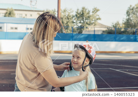 woman helps girl put on helmet at skating rink. They are engaged in bonding activity, enjoying quality time together outdoors woman helps girl put on helmet at skating rink. They are engaged in bonding activity, enjoying quality time together outdoors 140055323