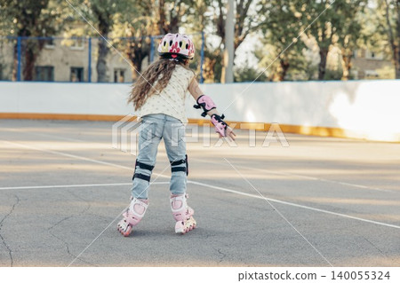 young Caucasian girl with long brown hair skates on a smooth surface. She wears a pink helmet and protective gear, enjoying quality time outdoors. Back view young Caucasian girl with long brown hair skates on a smooth surface. She wears a pink helmet and protective gear, enjoying quality time outdoors. Back view 140055324