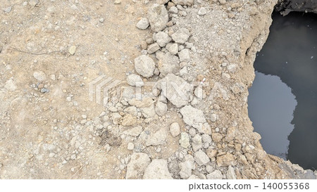 An extreme close-up of a construction or ground site showing light brown dirt gravel sloping down to a dark, water-filled pit. The edge is lined with loose rocks clumps of earth. An extreme close-up of a construction or ground site showing light brown dirt gravel sloping down to a dark, water-filled pit. The edge is lined with loose rocks clumps of earth. 140055368