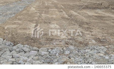 A construction scene with large gray rocks gravel in the foreground. The background is compacted brown earth soil displaying clear tire tracks from heavy machinery. A construction scene with large gray rocks gravel in the foreground. The background is compacted brown earth soil displaying clear tire tracks from heavy machinery. 140055375