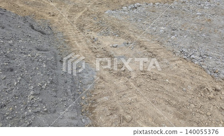 The image shows a construction site with gray gravel on the left right of a central path of light brown dirt. Tire marks are visible on the disturbed earth. The image shows a construction site with gray gravel on the left right of a central path of light brown dirt. Tire marks are visible on the disturbed earth. 140055376
