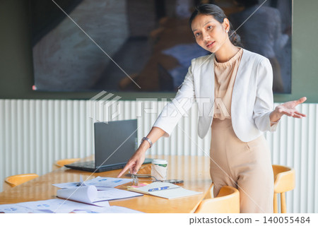 Professional businesswoman pointing to sales documents on office table with laptop, coffee, and notebook, explaining project details, strategy planning, business review, and team discussion. 140055484
