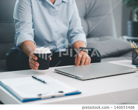 Professional reaching for closed laptop while holding takeaway coffee cup at modern office desk, showing business break, remote work routine, planning, productivity, and clean workspace. Professional reaching for closed laptop while holding takeaway coffee cup at modern office desk, showing business break, remote work routine, planning, productivity, and clean workspace. 140055486