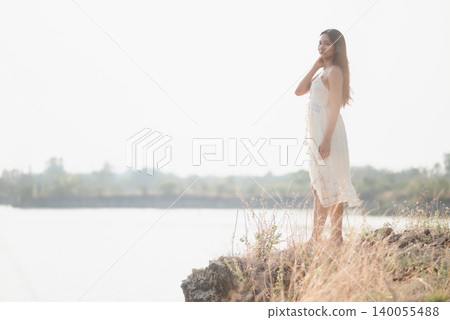 Serene woman in white lace dress standing on rocky lakeside cliff in soft morning light, gazing calmly across water, showing peaceful travel, summer nature, freedom, and wellness lifestyle. Serene woman in white lace dress standing on rocky lakeside cliff in soft morning light, gazing calmly across water, showing peaceful travel, summer nature, freedom, and wellness lifestyle. 140055488