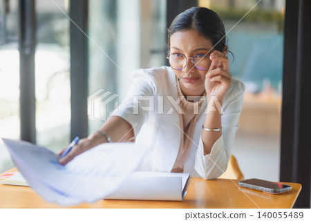 Serious businesswoman reaching across office table with documents and pen, reviewing report details, discussing data, business decision, problem solving, and focused professional planning. Serious businesswoman reaching across office table with documents and pen, reviewing report details, discussing data, business decision, problem solving, and focused professional planning. 140055489