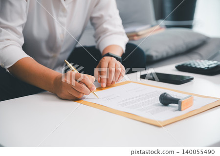 Hand signing business document on folder with stamp, smartphone, and calculator nearby, showing contract approval, legal paperwork, finance review, and office administration workflow. Hand signing business document on folder with stamp, smartphone, and calculator nearby, showing contract approval, legal paperwork, finance review, and office administration workflow. 140055490