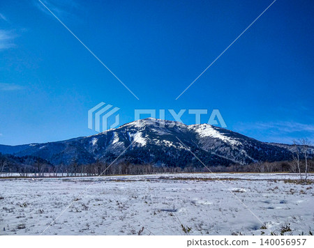 A stunning photograph of snow-covered Mt. Shibutsu viewed from Oze Marsh in early winter, November, against a clear blue sky and vast snowfields. 140056957