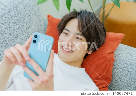 A young Japanese man is lying on a sofa looking at his smartphone. A young Japanese man is lying on a sofa looking at his smartphone. 140057471