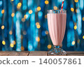 Strawberry milkshake in tall glass with striped straw on rustic wooden table, set against vibrant blue backdrop with golden bokeh for festive, refreshing scene 140057861