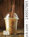 Iced coffee in clear plastic takeaway cup with ice and brown straw, condensation beading on the exterior, resting on rustic wooden table for refreshing summer drink 140057865