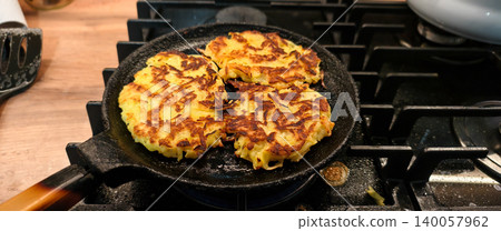 Crispy golden brown potato pancakes frying in a hot cast iron pan on a gas stove burner, preparing a delicious homemade meal or snack in a kitchen setting. Crispy golden brown potato pancakes frying in a hot cast iron pan on a gas stove burner, preparing a delicious homemade meal or snack in a kitchen setting. 140057962