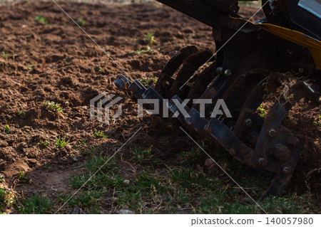 Walk-behind tractor tilling garden soil with rugged blades, preparing earth for planting and new crops; close-up of rototiller working rich brown soil in spring sunlight.. Walk-behind tractor tilling garden soil with rugged blades, preparing earth for planting and new crops; close-up of rototiller working rich brown soil in spring sunlight.. 140057980