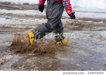 Child wearing colorful waterproof clothing and yellow rubber boots jumping and splashing in a dirty, muddy puddle outdoors on a wet, snowy day, creating dynamic motion and joyful play. 140058004