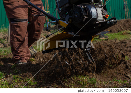 Man operating a walk-behind tractor to plow and cultivate garden soil, preparing rows for spring planting on a small farm or home garden in rural outdoors. Man operating a walk-behind tractor to plow and cultivate garden soil, preparing rows for spring planting on a small farm or home garden in rural outdoors. 140058007