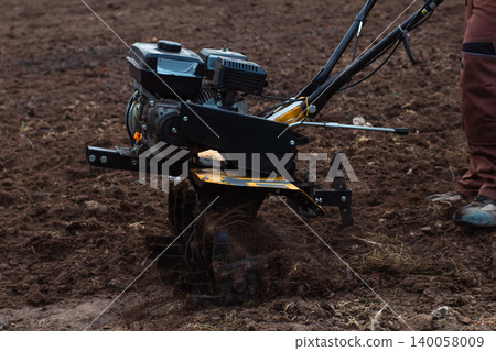 Man operating a walk-behind tractor, cultivating and preparing the soil for planting in a home garden, demonstrating spring gardening work and agricultural practices. Man operating a walk-behind tractor, cultivating and preparing the soil for planting in a home garden, demonstrating spring gardening work and agricultural practices. 140058009