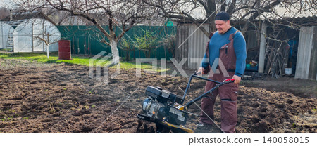 Farmer using a motorized walk-behind tractor to till and prepare garden soil for spring planting near greenhouses in a rural backyard small-farm setting, daytime work. Farmer using a motorized walk-behind tractor to till and prepare garden soil for spring planting near greenhouses in a rural backyard small-farm setting, daytime work. 140058015