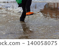 Child wearing black pants and bright orange waterproof boots jumping over a slushy puddle, splashing water and melting snow on a mild winter or early spring day. 140058075