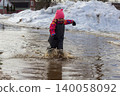 Little girl wearing warm clothes and a pink hat joyfully splashing in a large muddy puddle formed by melting snow, embodying the innocence and fun of childhood during early spring. 140058092
