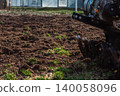 Walk-behind tractor tilling brown earth in a home garden, preparing the ground for planting season, with fresh green grass emerging and greenhouses in the background. 140058096