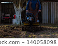 Man operating a walk-behind tractor tilling rich black soil in a home garden, preparing ground for planting and cultivating crops during spring seasonal farm work. 140058098