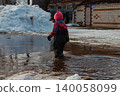 Child wearing winter clothes and a pink hat playing in a large muddy puddle, surrounded by melting snow and slush, enjoying a playful outdoor activity during a sunny winter day. 140058099