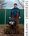 Farmer in work clothes operating a walk-behind tractor, cultivating and preparing the brown soil of a home garden or field for planting crops in a rural outdoor setting. 140058101