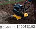 Man holding the handle of a gasoline-powered walk-behind tractor, cultivating fresh brown earth in a backyard garden plot on a spring day, preparing for planting. 140058102