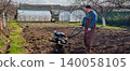 Man working in the garden, operating a walk-behind tractor to cultivate the soil. Greenhouses are visible in the background, showing a scene of active farm work and preparing the ground for planting. 140058105