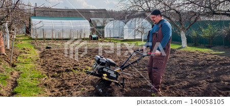 Man working in the garden, operating a walk-behind tractor to cultivate the soil. Greenhouses are visible in the background, showing a scene of active farm work and preparing the ground for planting. Man working in the garden, operating a walk-behind tractor to cultivate the soil. Greenhouses are visible in the background, showing a scene of active farm work and preparing the ground for planting. 140058105