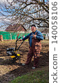 Man in workwear standing by a walk-behind tractor on freshly tilled garden soil, preparing planting beds for spring crops in a sunny, rural home-garden setting. 140058106