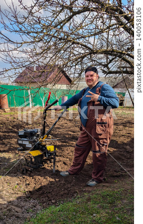 Man in workwear standing by a walk-behind tractor on freshly tilled garden soil, preparing planting beds for spring crops in a sunny, rural home-garden setting. Man in workwear standing by a walk-behind tractor on freshly tilled garden soil, preparing planting beds for spring crops in a sunny, rural home-garden setting. 140058106