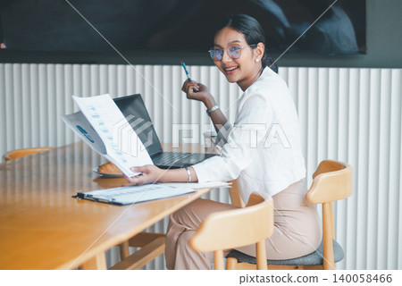 Smiling businesswoman holding report beside laptop at office table, creating a friendly workplace mood with documents, coffee and pastry during a positive business discussion session. Smiling businesswoman holding report beside laptop at office table, creating a friendly workplace mood with documents, coffee and pastry during a positive business discussion session. 140058466