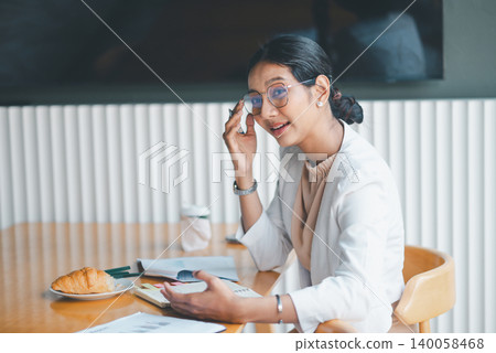Thoughtful businesswoman touching glasses while talking at office desk, reviewing notes and documents beside coffee and pastry during a professional morning discussion session. Thoughtful businesswoman touching glasses while talking at office desk, reviewing notes and documents beside coffee and pastry during a professional morning discussion session. 140058468