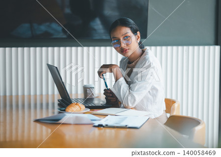 Thoughtful businesswoman working with laptop, coffee and pastry at office table, reviewing documents and planning tasks in a calm professional workspace during a productive morning. Thoughtful businesswoman working with laptop, coffee and pastry at office table, reviewing documents and planning tasks in a calm professional workspace during a productive morning. 140058469