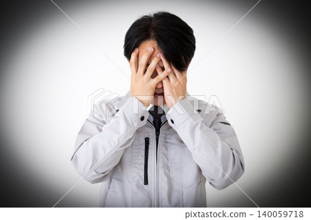 A middle-aged businessman in gray work clothes, covering his face and looking dejected, against a white background. A middle-aged businessman in gray work clothes, covering his face and looking dejected, against a white background. 140059718